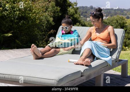 Woman and child sitting on cushioned lounge on wood deck in towels applying sunscreen Stock Photo