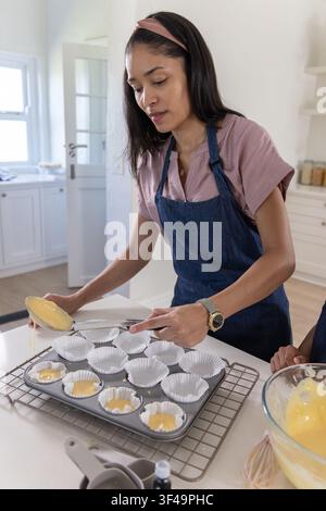 African-American mom in denim apron scooping batter into muffin tin as youth holds bowl in kitchen Stock Photo
