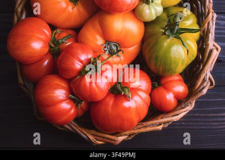 Overhead view of mixed heirloom tomatoes in wicker basket. Seasonal organic harvest, rustic kitchen mood Stock Photo