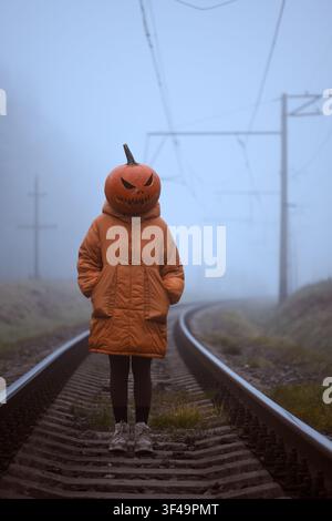 Pumpkin headed figure standing alone on railway tracks disappearing into dense fog. Surreal Halloween concept with abandoned railroad and eerie autumn atmosphere Stock Photo