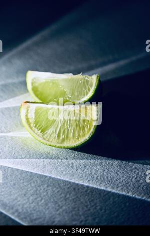 Close-up photo of two fresh, juicy lime wedges illuminated by a strong spot of light creating ray shadows on a textured dark surface. Modern creative food photography featuring citrus fruit, vibrant green pulp, hard light contrast, and moody atmosphere. Ideal for bar menus, cocktail concept visuals, and gourmet culinary projects. Stock Photo