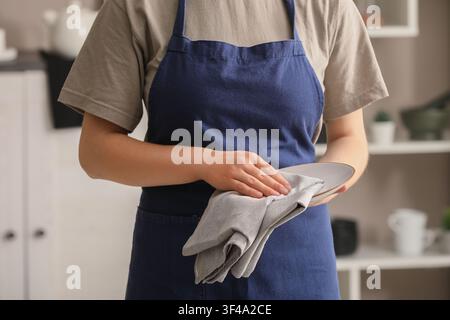 Person in a blue apron drying a clean plate with a gray cloth in a modern kitchen setting, Moment of domestic routine and attention to cleanliness in Stock Photo