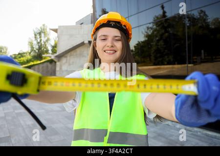 Smiling female construction worker in a hard hat and safety vest holding a measuring tape outdoors, the woman is focused on the measurement, precision Stock Photo