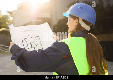 Female architect in a blue hard hat and safety vest reviewing a guest house plan at a construction site, She holds the architectural drawing with both Stock Photo