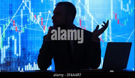 Silhouette of a businessman with a laptop in front of a blue screen displaying financial stock market data Stock Photo