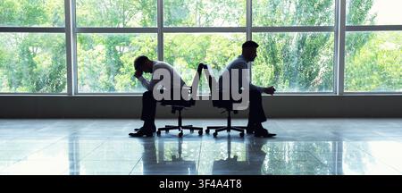 Two men in silhouette sitting back to back in office chairs conflict disagreement and relationship problems, the other man sits with clasped hands app Stock Photo