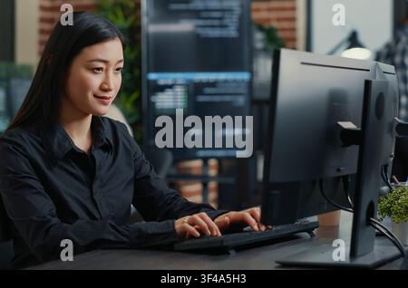 Smiling Asian Programmer Writing Code at Desk at a software development desk after compiling source code successfully, showing algorithm work and coding success. Stock Photo