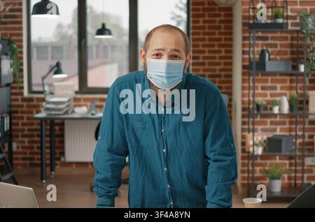 Masked businessman plans project strategy while reviewing charts, laptop data and project documents with protective face mask use in a startup office. Stock Photo