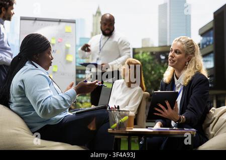 Diverse coworkers exchanging ideas in informal outdoor meeting, strategy workshop with women checking notes on tablet and laptop. Business partnership in daylight. Stock Photo