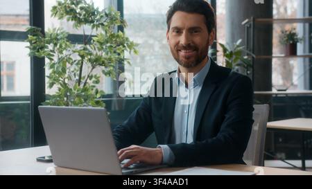 Caucasian businessman typing laptop in office looking at camera smiling posing portrait business man male guy happy entrepreneur workplace Stock Photo