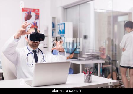 Young physician using virtual reality goggles in hospital office, with disabled senior woman in wheelchair talking with female doctor on clinic corridor, nurse in blue uniform holding patient x-ray. Stock Photo