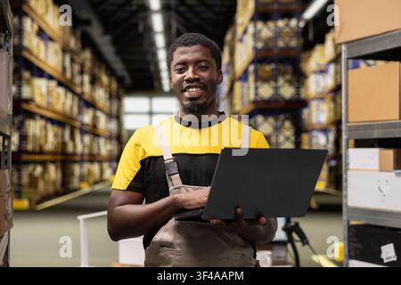 Portrait of smiling black man in workwear checking cargo parcels on laptop inventory system, managing fulfillment center operations for efficient distribution. E-commerce large business. Stock Photo