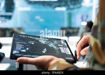 Close up of military specialist using tablet displaying aerial tactical map. Army operator selects targets on device touchscreen while monitoring command center surveillance Stock Photo