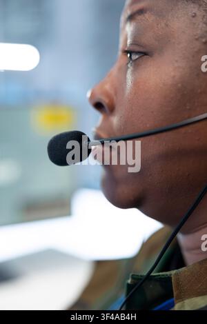 Close up of military operator speaking through headset microphone while monitoring control room. Female army dispatcher relays mission updates to personnel from command center Stock Photo