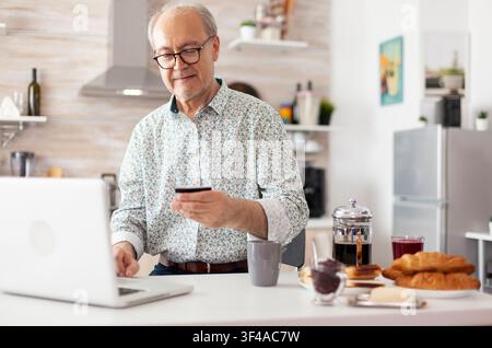 Senior man completes an online payment on a laptop during breakfast in the kitchen, using banking technology and internet shopping tools as part of a natural digital communication moment involving. Stock Photo