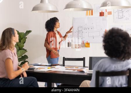 Diverse female coworkers presenting plans and reviewing swatches at modern studio table with laptop. Design, architecture, collaboration, teamwork, blueprints, palette, interior Stock Photo