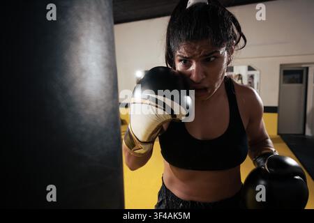 Female athlete trains intensely on a heavy punching bag in a gym, showing determination, focus, sweat and strength during a powerful boxing workout and conditioning session. Stock Photo
