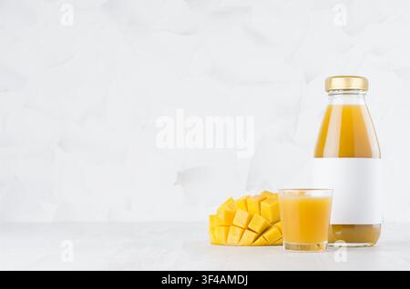 Bright orange pulpy mango juice in glass bottle mock up with blank label, wine glass, fruit pieces on white wood table in light interior, template for packaging, advertising, design, branding product. Stock Photo