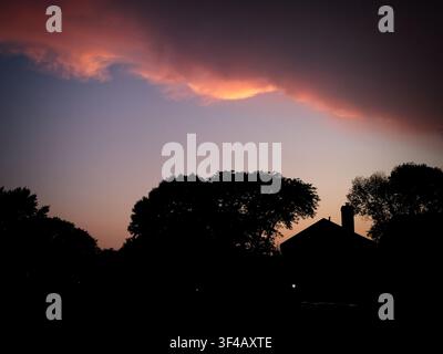 Vibrant pink and orange sunset sky with dramatic dark storm clouds casting a silhouette over trees and houses Stock Photo