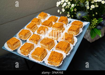 Square pastries with golden topping arranged neatly on a blue tray. White paper liners beneath each pastry. Fresh flower arrangement beside the tray. Stock Photo