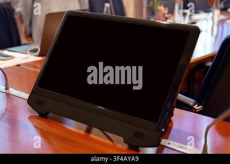Black monitor on wooden table. Screen dark, no visible content. Modern device sits in office setting. Background shows blurred chairs and desks. Suita Stock Photo