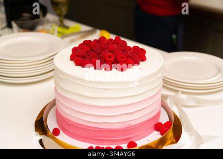 A beautifully frosted ombre cake featuring gradients of pink and white, topped generously with fresh raspberries. The cake is set on a white table sur Stock Photo