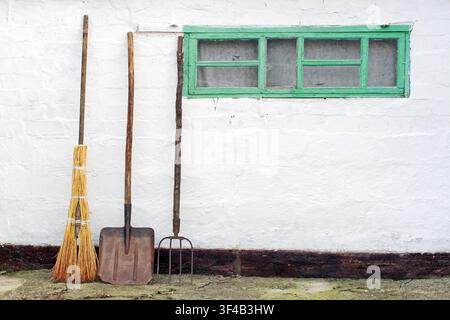 Old garden tools near the wall of the barn. A broom, a shovel and a pitchfork against the wall with a window. Stock Photo