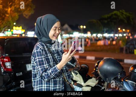 Asian Indonesian woman checking her smartphone near a busy night market in Yogyakarta. Mobile technology and modern urban lifestyle at night. Stock Photo