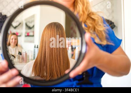 Hairdresser holding a mirror, showing long brown hair to a smiling client in a salon Stock Photo