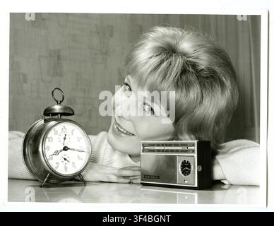Transistor radio, with clock, versus alarm clock, advertising photography, smiling woman leaning at table with alarm clock and retro radio in a black Stock Photo