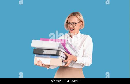 Stressed female secretary overloaded with paperwork, holding folders with documents on blue background Stock Photo