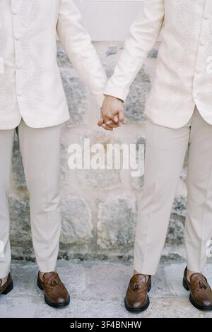 Partners in Matching Cream Suits Holding Hands Together by Rustic Stone Wall, Matching Beige Trousers and Brown Shoes, Subtle Ring Glimpses Stock Photo