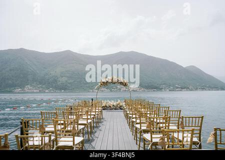 A beautifully arranged outdoor wedding ceremony setup on a wooden pier overlooking the Tranquil Bay and Lush Green Mountains of Montenegro. Rows of el Stock Photo