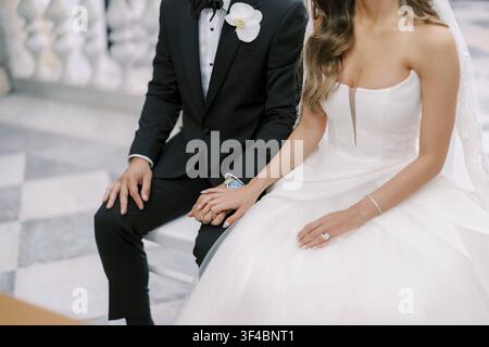 Bride And Groom Seated Portrait With Hands Linked, Groom In Tuxedo And Bride In Strapless Gown, Soft Indoor Light On Formal Attire, Detail Stock Photo