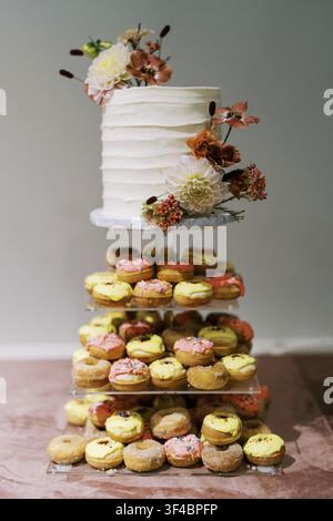 Tiered cake and donut tower showcasing white frosted cake with cascading floral arrangement, pastel glazed donuts stacked on wooden stand Stock Photo