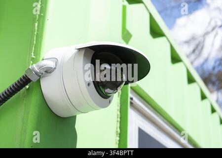 A white dome security camera is mounted on a bright green wall, with a reflection visible in the camera lens. The camera is connected by a black condu Stock Photo