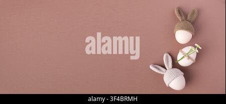 Three decorative Easter eggs wearing knitted bunny hats, one adorned with a flower, arranged on a soft pink background for festive decoration Stock Photo