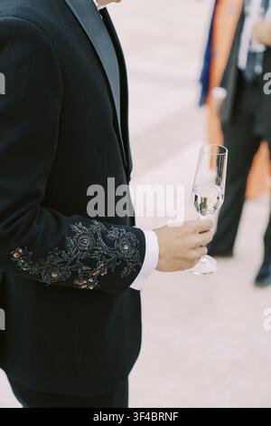 A man dressed in a black suit with intricate floral embroidery on the sleeve holds a glass of champagne at a formal event. Another person in formal at Stock Photo