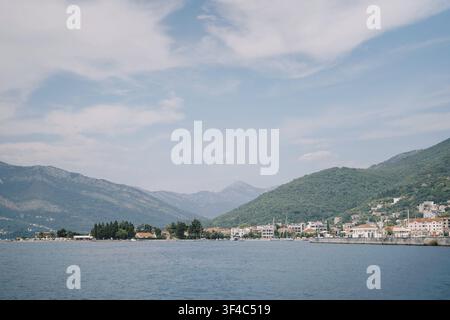 Calm Sea, Mountains Framing Coastal Village. Soft Blue Sky With Scattered Clouds, Distant Quay With Redroof Houses Stock Photo