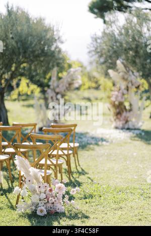 Outdoor aisle lined with wooden chairs and pastel floral clusters, olive trees and sunlit bokeh in background, bouquet marker at aisle end, planner ar Stock Photo