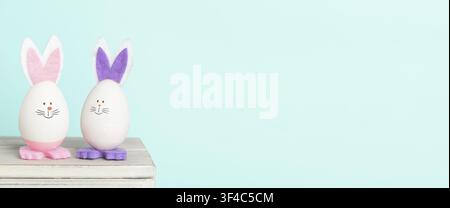 Two decorative Easter eggs with bunny ears sit on a wooden surface, featuring pink and purple accents against a light blue background Stock Photo