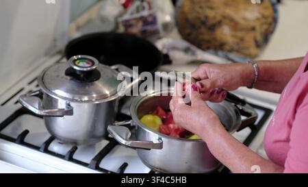 Mature hands add cut tomato to colorful peppers in a metal pot, shown in a tight side composition. The realistic image connects homemade meals with Stock Photo