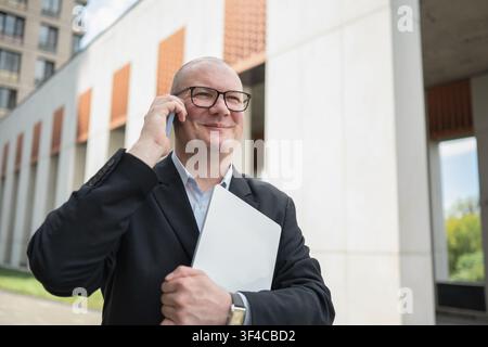 Happy mature businessman talking on smartphone while carrying laptop outside modern office building. Business communication, leadership, success and p Stock Photo