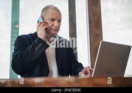 Serious mature businessman talking on smartphone while working on laptop in modern workspace. Business communication, multitasking and remote work con Stock Photo