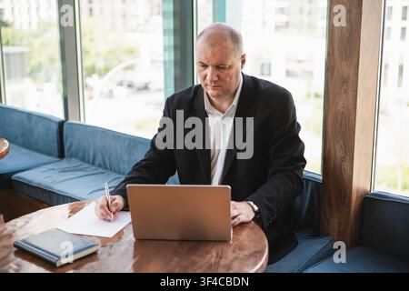 Focused mature businessman writing notes while using laptop in modern cafe workspace. Business planning, productivity and remote office concept. Stock Photo