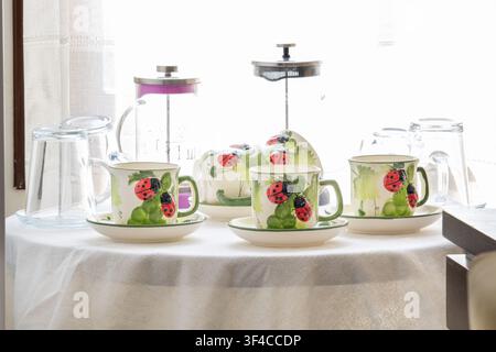 A charming tea set featuring ceramic cups and saucers decorated with hand-painted ladybug and leaf motifs, neatly arranged on a white tablecloth Stock Photo