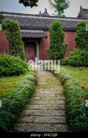 A textured stone path, lined with vibrant green grass, leads directly towards a red wall building featuring a dark gray tiled roof and an arched entry Stock Photo