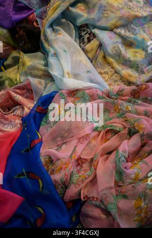 A vibrant pile of loosely folded sheer fabrics fills the frame, showcasing various patterns and colors. Visible textiles include a bright blue fabric Stock Photo