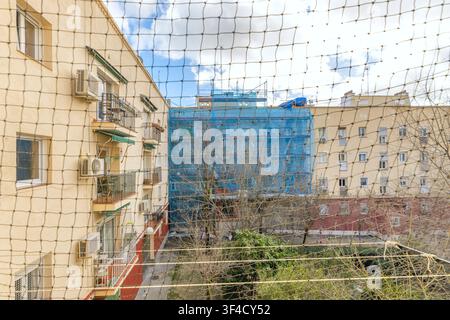 Interior courtyard view through a protective net, showing apartment balconies, air conditioners, and a scaffolded building under renovation. Stock Photo