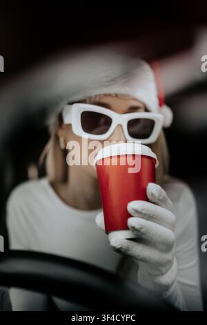 Woman wearing santa hat and sunglasses drinking from a red cup inside a car during winter Stock Photo